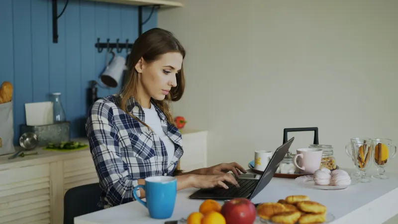 Een volwassen vrouw zit rustig aan de keukentafel met haar laptop